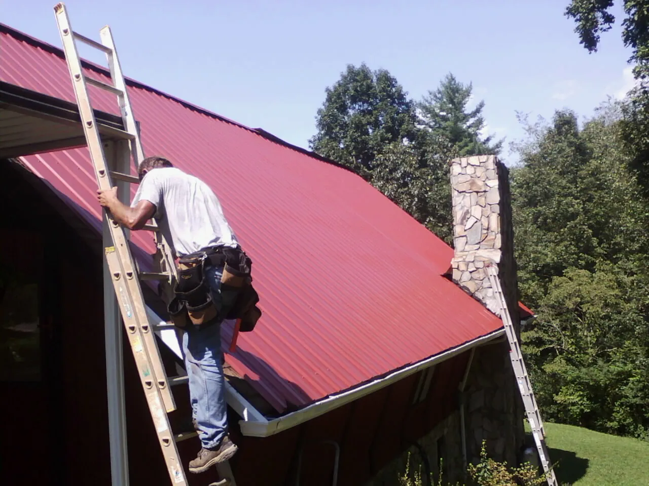 Roofer on a ladder beside a red metal roof and stone chimney — Soffit & Fascia Repair by Roofing Brothers in Frisco